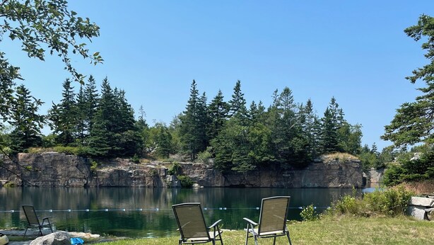 A grassy area with two chairs facing a calm lake, surrounded by trees and rocky cliffs. - The Bodwell Cottage at East Boston Quarry (Vinalhaven)