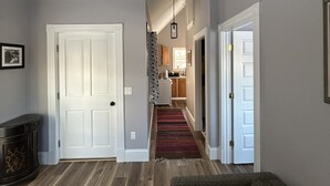 A hallway with a wooden floor, a red and black striped rug, a gray sofa, and a kitchen visible in the background. - The Bodwell Cottage at East Boston Quarry (Vinalhaven)