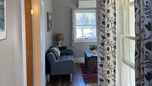 A hallway with a grey sofa, a patterned rug, and a window with patterned curtains. - The Bodwell Cottage at East Boston Quarry (Vinalhaven)