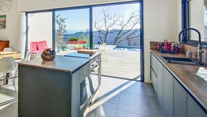 Eine moderne Küche mit einer großen Kochinsel, einem Essbereich mit einem rosa Stuhl sowie Blick auf eine Terrasse und Berge durch eine Glastür.
