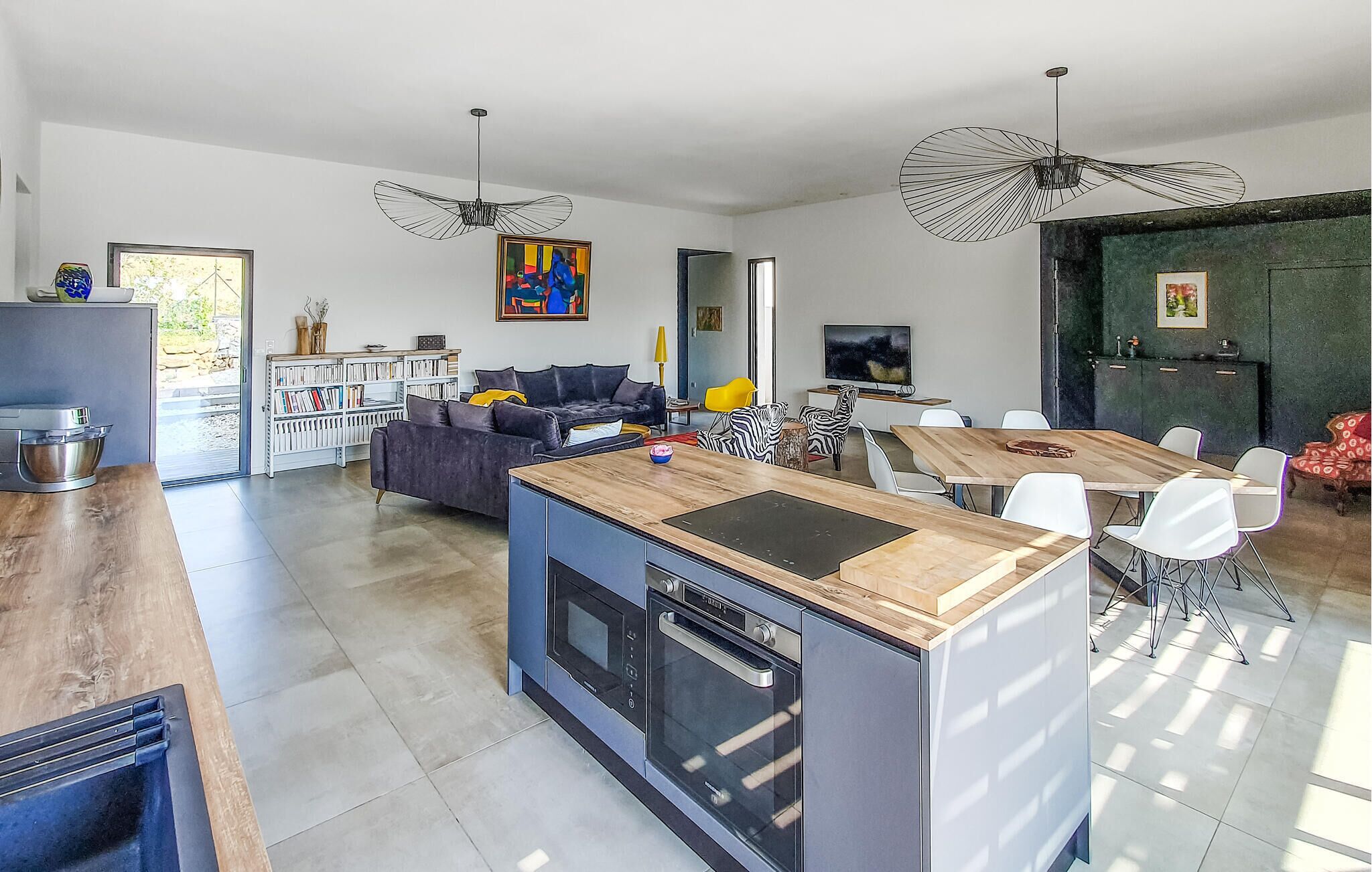 Uma cozinha moderna com uma ilha central, uma sala de jantar com mesa de madeira e uma sala de estar com sofá e pintura.