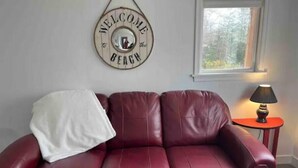 A red leather sofa with a white throw blanket, a wall-mounted welcome sign, a small table with a lamp, and a window with a view of trees.