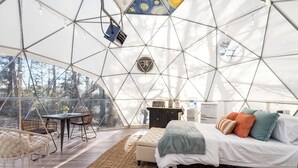 Ein geräumiges Zimmer mit einem großen Bett, einem kleinen Tisch und Stühlen, mit geometrischer Decke und großen Fenstern.