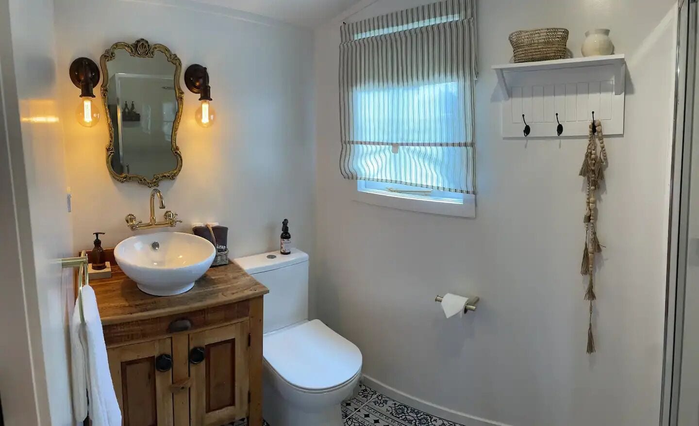 A bathroom with a wooden vanity, white sink, and a large mirror. There is a toilet and a window with curtains.