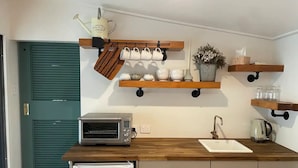 Cocina compacta con tostadora, pileta y heladera pequeña. Hay repisas de madera sobre la mesada con tazas y una regadera.