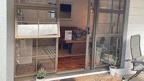 Una habitación con un amplio ventanal corredizo, piso de madera, una pequeña planta en maceta, una silla de mimbre y una mesa con planta.