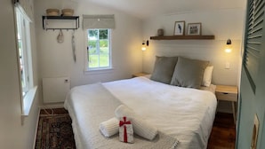 Una cama bien hecha con edredón blanco y almohadas, una mesita de noche con lámpara, una ventana con cortinas y un estante montado en la pared con objetos decorativos.