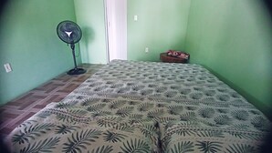 Una stanza con un letto, una ventola da terra, una porta e un piccolo tavolo con un oggetto decorativo.