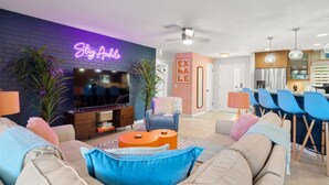 Una sala de estar moderna con un letrero de neón, un televisor de pantalla plana, un sofá, una mesa de centro y un comedor con sillas, con una cocina al fondo.