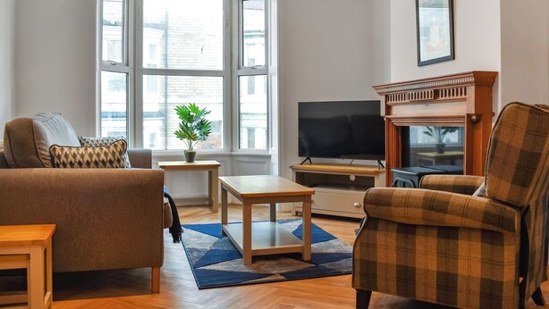 Een woonkamer met een bank, fauteuil, televisie, open haard en grote ramen.