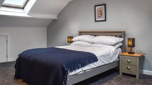 Een slaapkamer met dakraam, een bed met witte lakens, een blauwe plaid, een nachtkastje met lamp en een lijst met foto aan de muur.