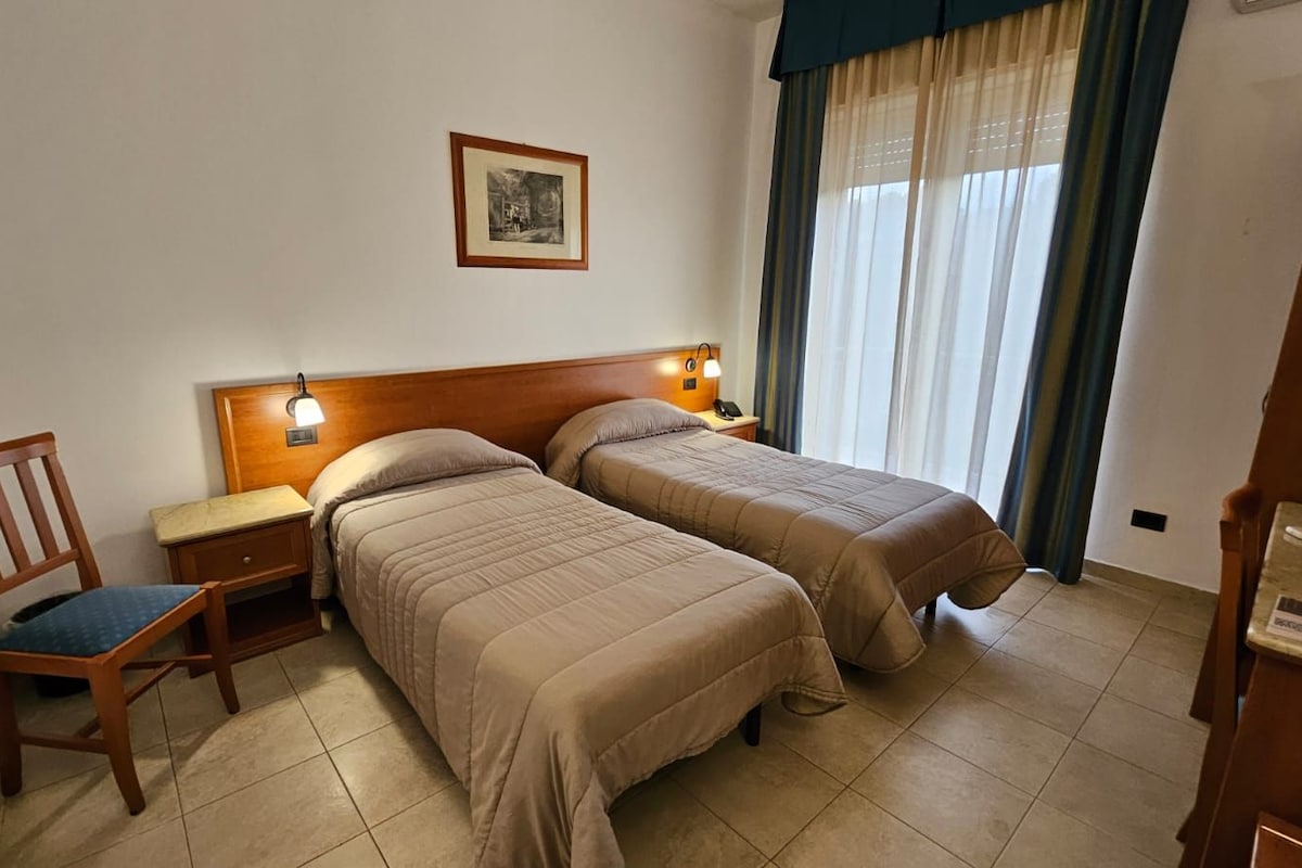 A hotel room with two beds, a chair, a nightstand, a lamp, and a picture on the wall.
