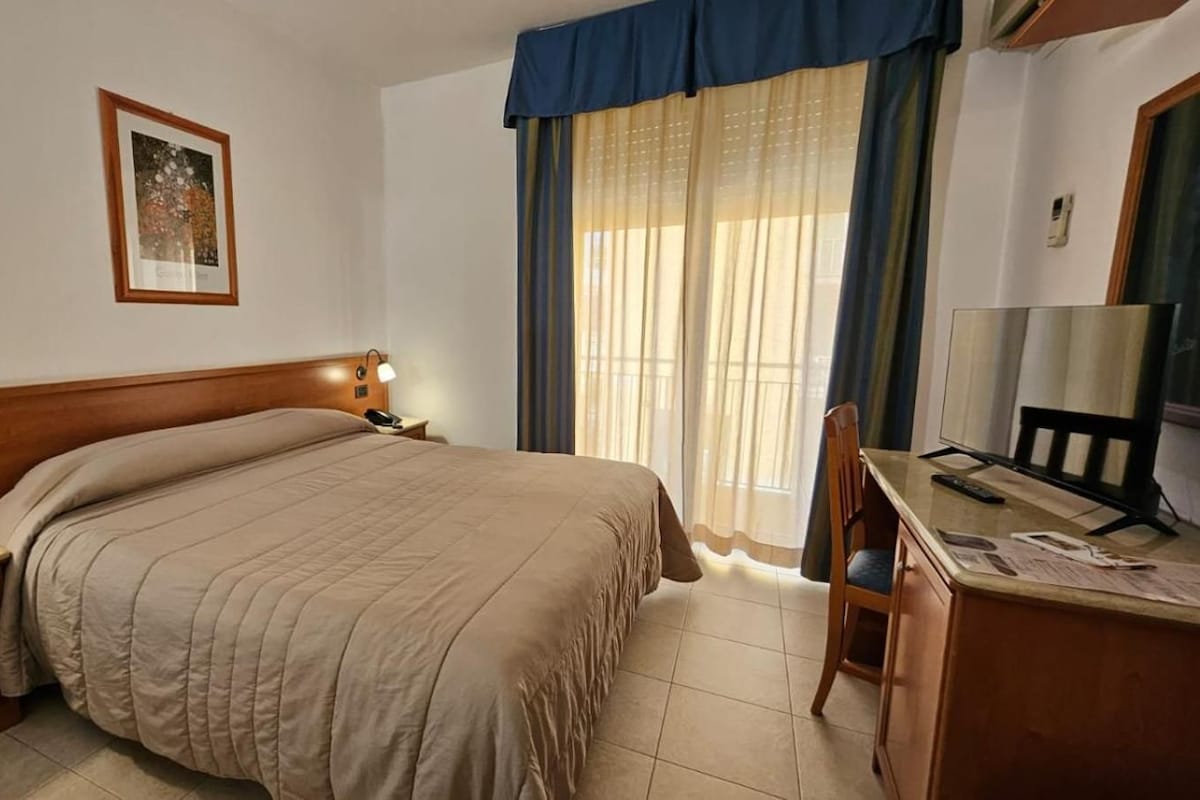 A hotel room with a bed, bedside table, lamp, desk with a chair, and a window with curtains.