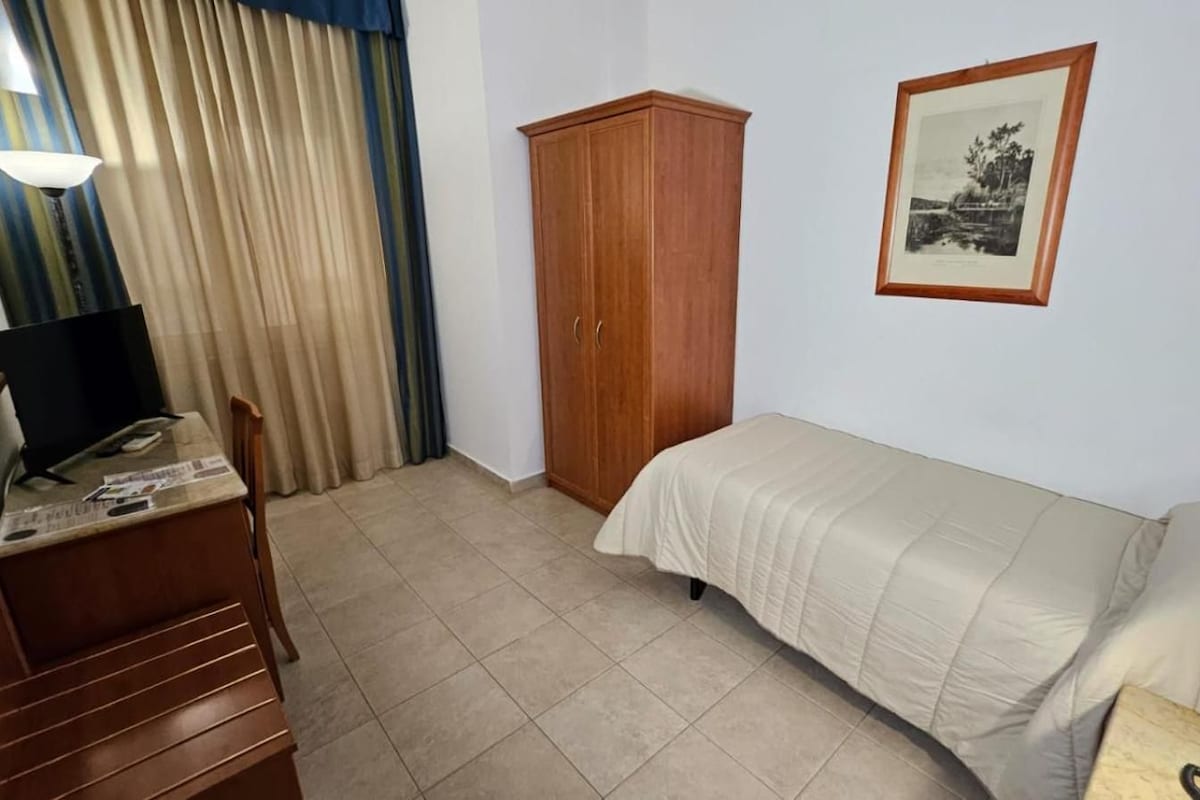 A bedroom with a bed, a wooden wardrobe, a desk with a computer, and a framed picture on the wall.