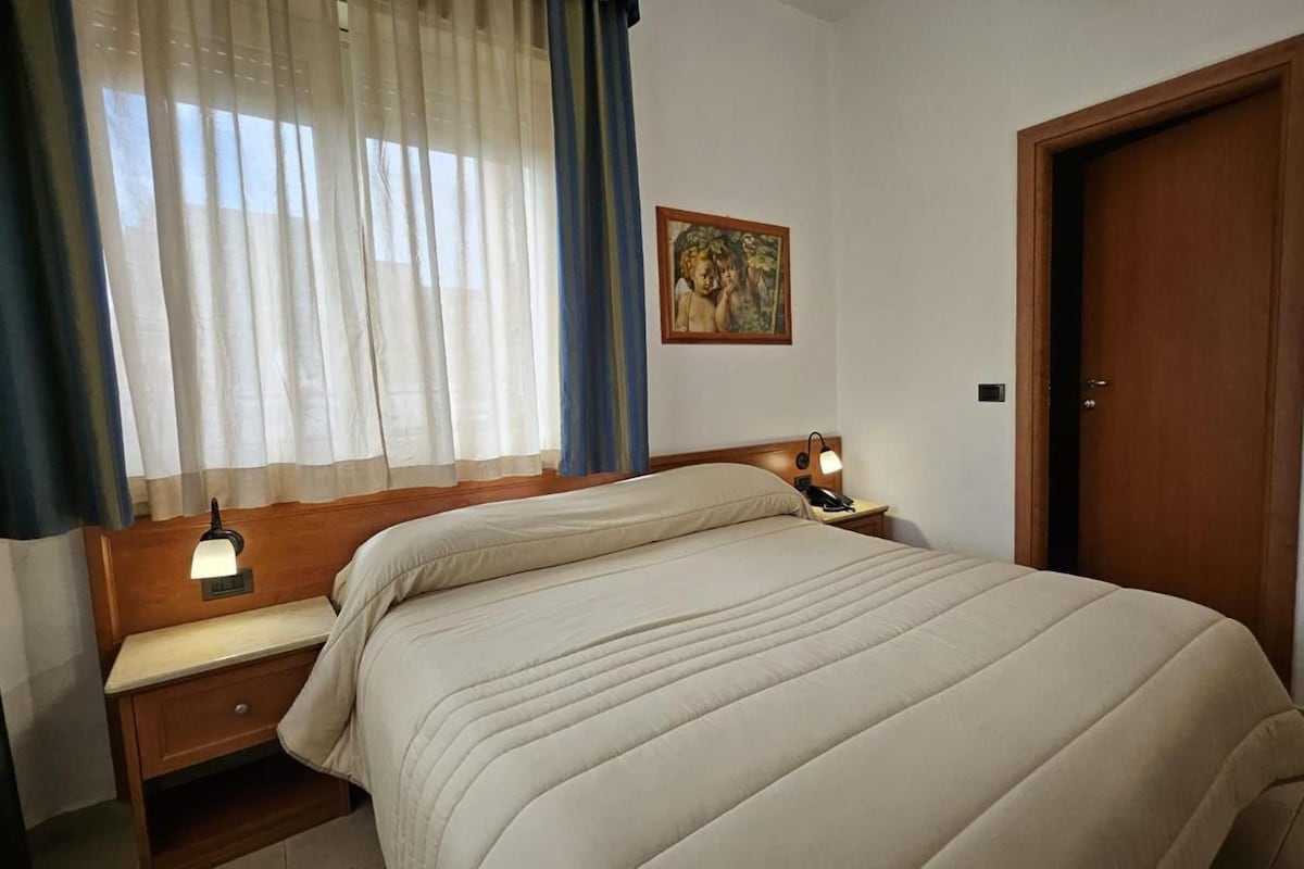 A hotel room with a bed, a bedside table with a lamp, a painting on the wall, and a door.