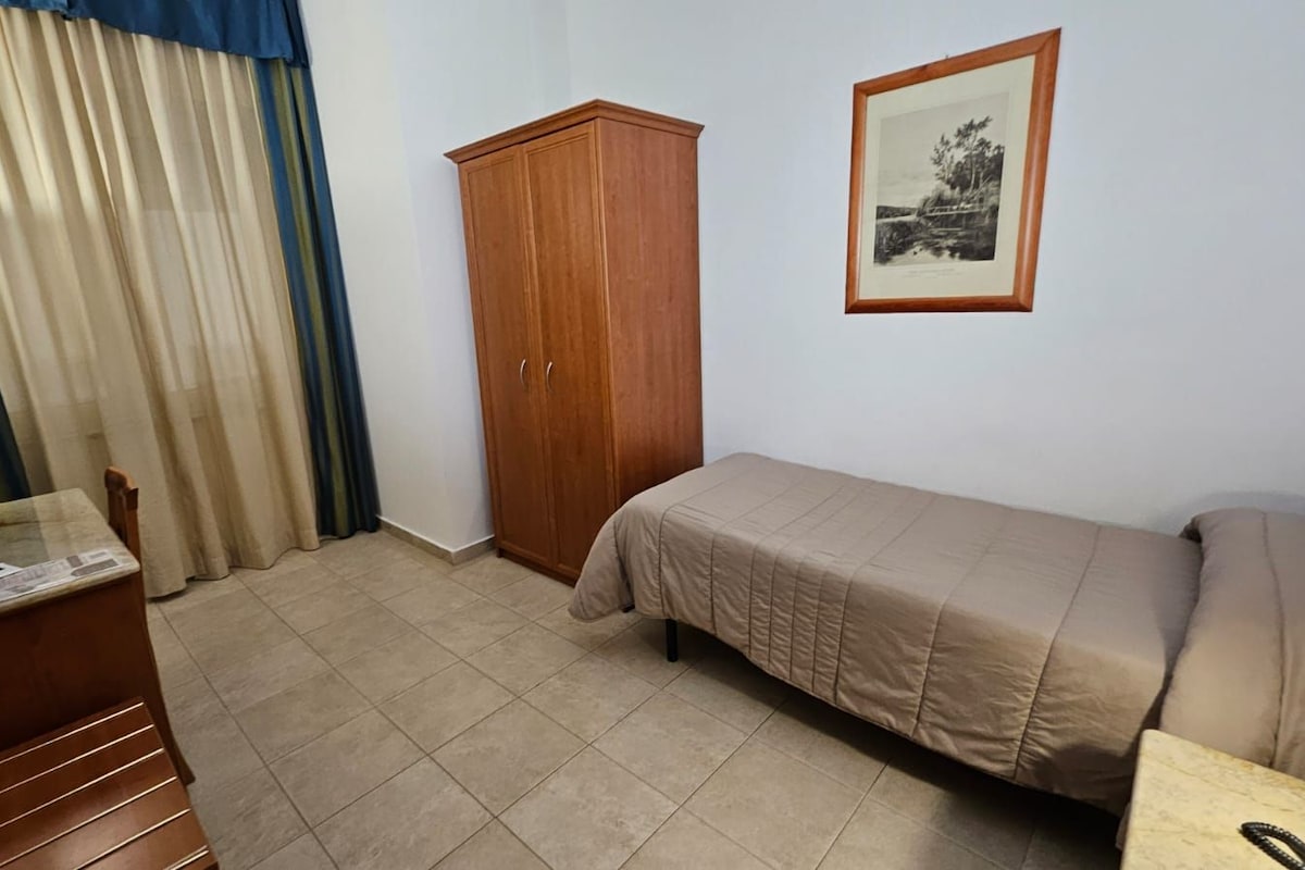 A small, tidy room with a bed, a wooden wardrobe, a desk with a lamp, and a framed picture on the wall.