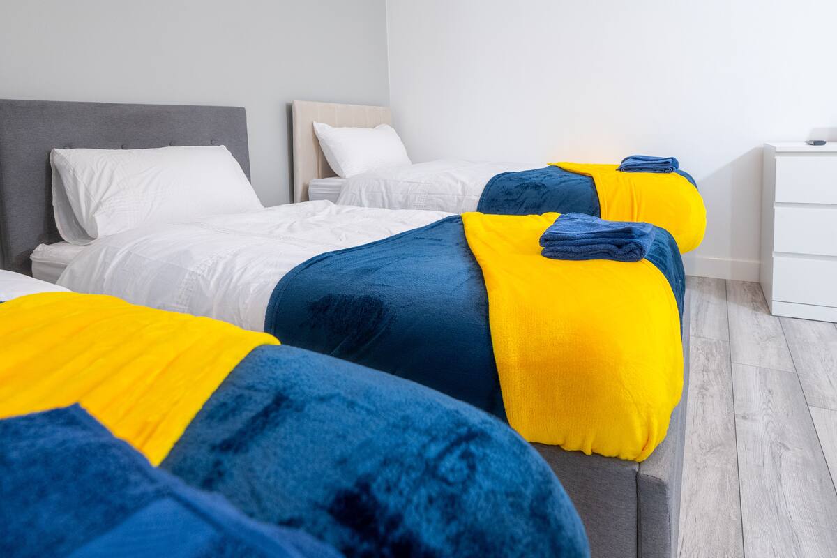 A hotel room with two beds, each with a grey headboard, white bedding, and blue and yellow throw pillows.