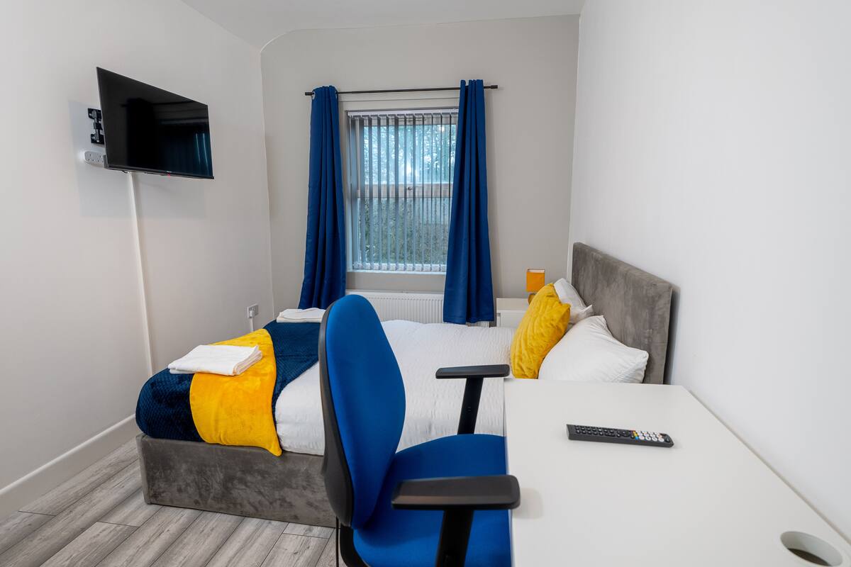 A hotel room with a bed, a TV mounted on the wall, a window with curtains, a bedside table, and a chair.