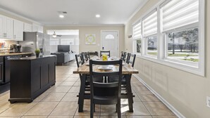 A modern kitchen with a central island, dining table, and chairs. There is a refrigerator, stove, and sink. The kitchen has large windows and a view of the outdoors. - Rising Tides Hideaway! (Long Beach)