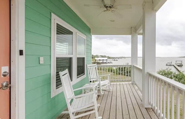 A porch with a view of the water, white chairs, and a green building. - Sunset Bay Pelican Cottage (Port Lavaca)