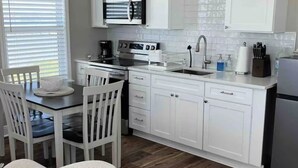 A modern kitchen with white cabinetry, a dining area with a table and chairs, and a wooden floor. - Sunset Bay Seagull Cottage (Port Lavaca)