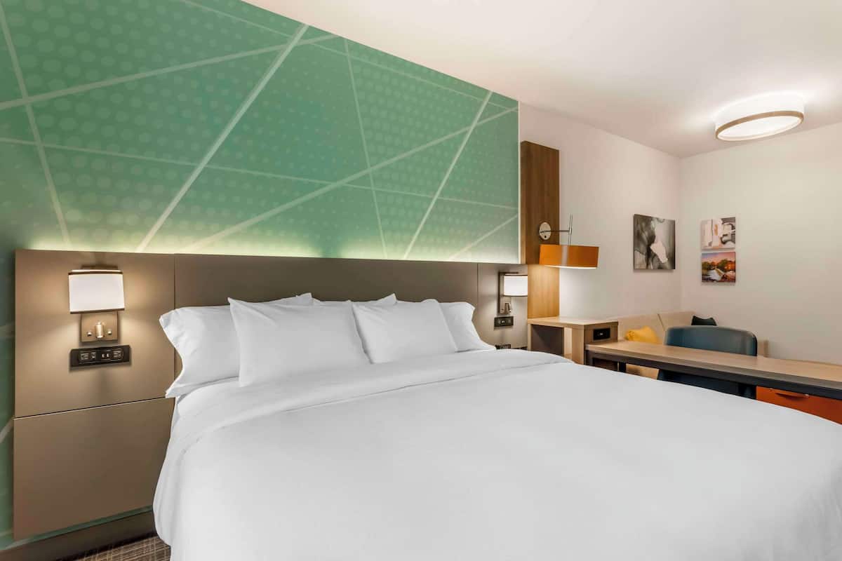 A hotel room with a large bed, a desk, a chair, a nightstand, and a wall-mounted lamp.