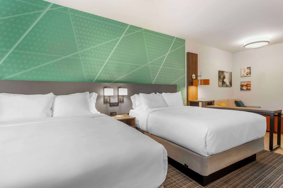 A hotel room with two beds, a green accent wall, a small desk with a lamp, and artwork on the wall.