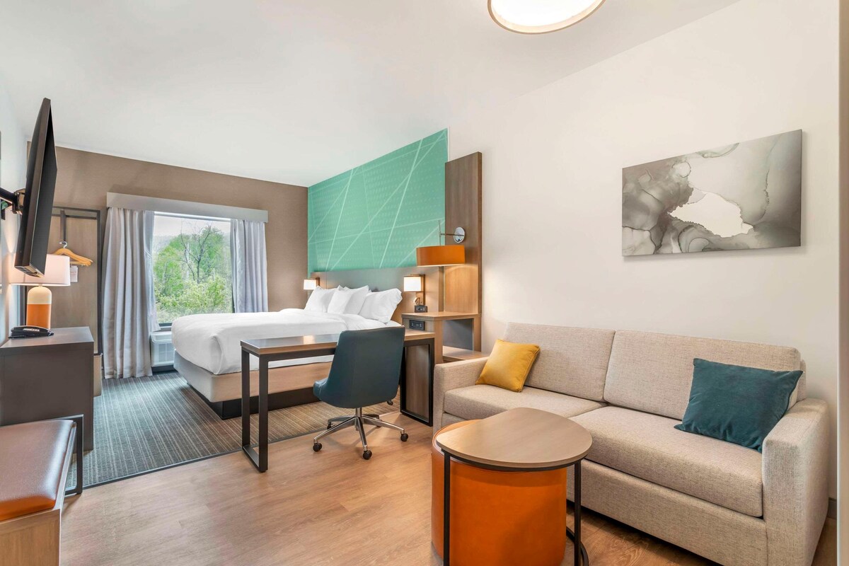 A hotel room with a bed, a sofa, a desk, a chair, a TV, and artwork on the walls.