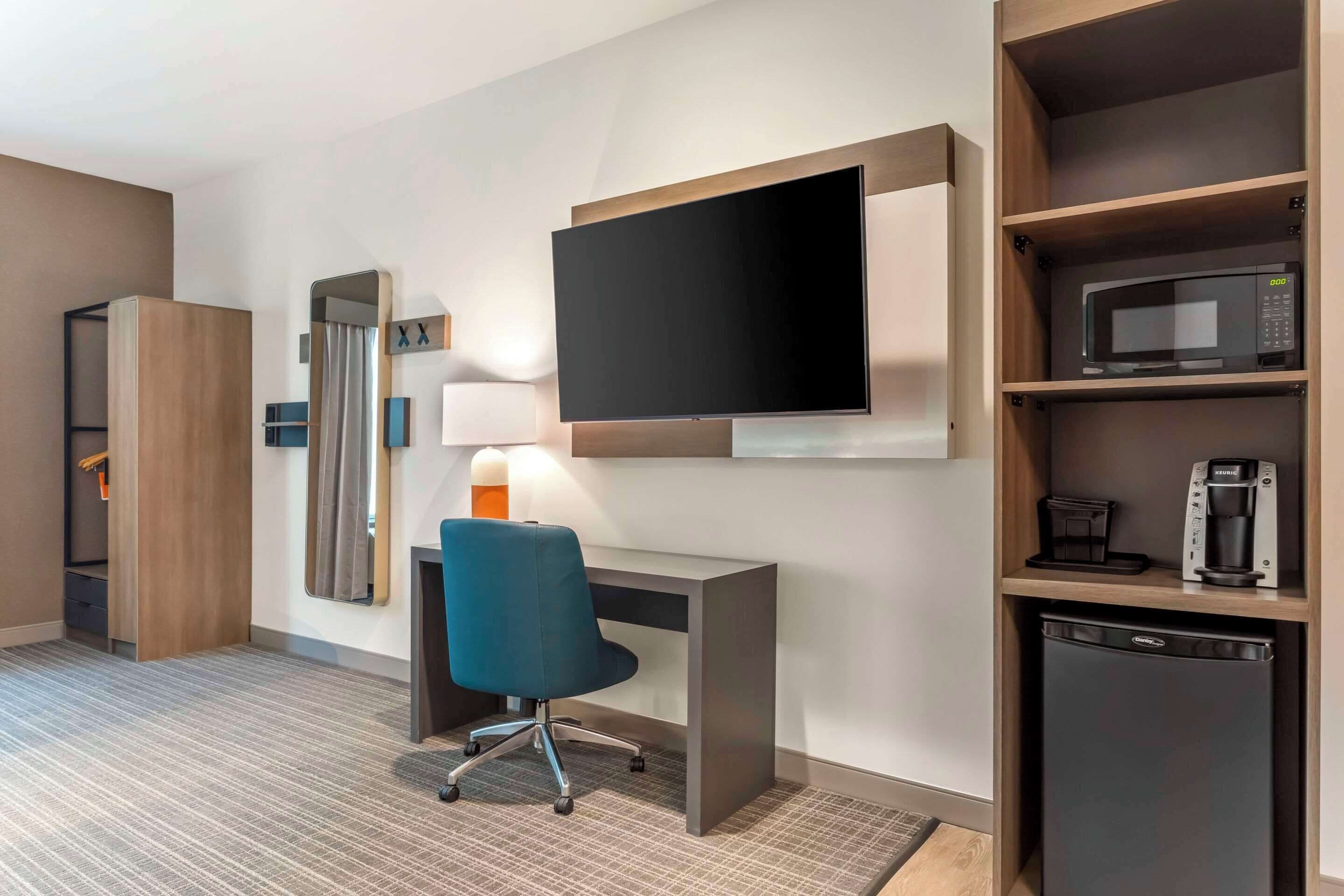 Habitación de hotel con televisión de pantalla plana montada en la pared, un escritorio con silla, microondas, cafetera y mini refrigerador.