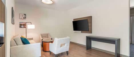 A modern living room with a sofa, armchair, round table, and a flat-screen TV mounted on the wall.