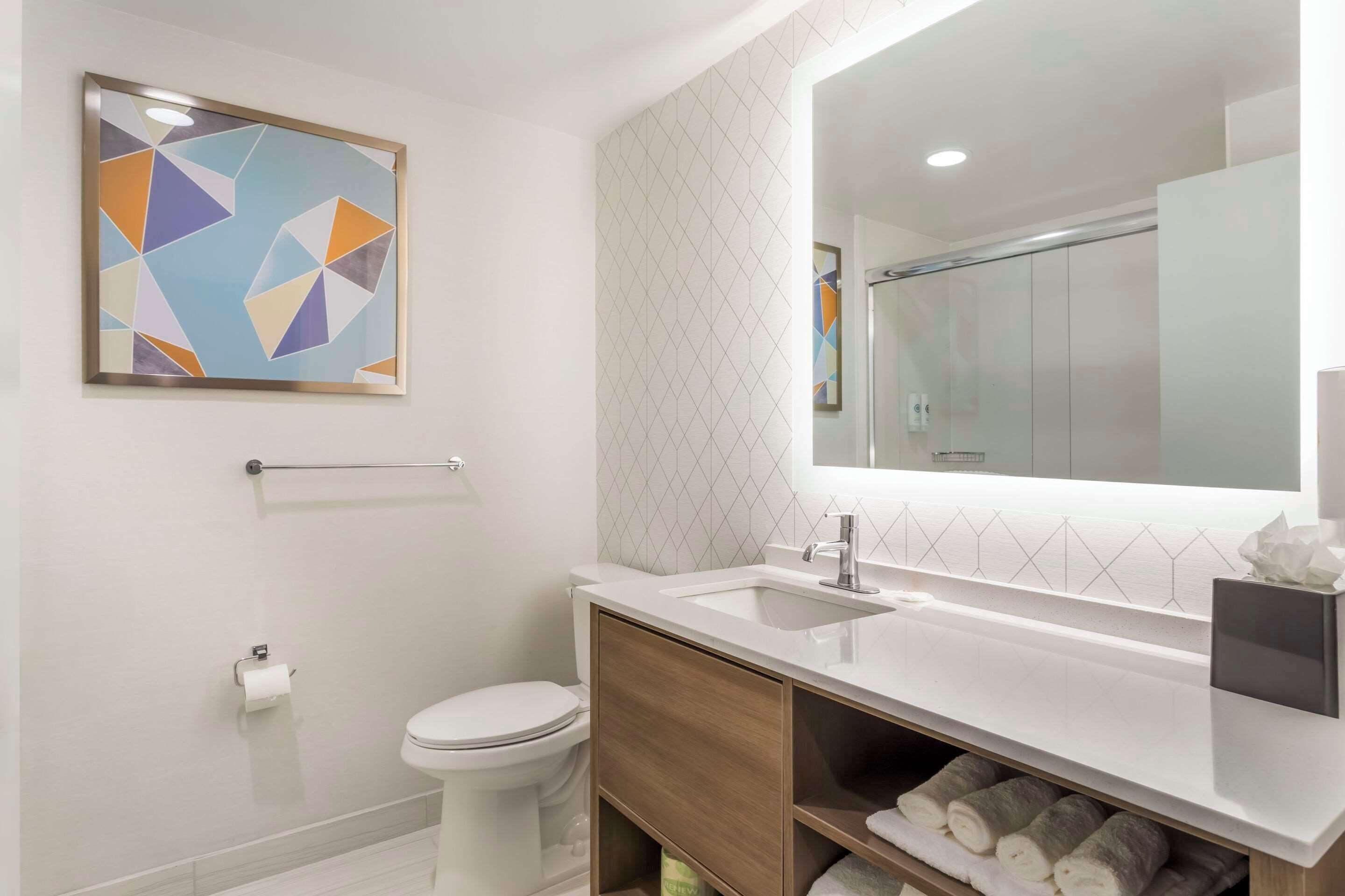 A modern bathroom with a white sink, mirror, and toilet. There is a towel rack above the toilet and a towel dispenser below the sink.