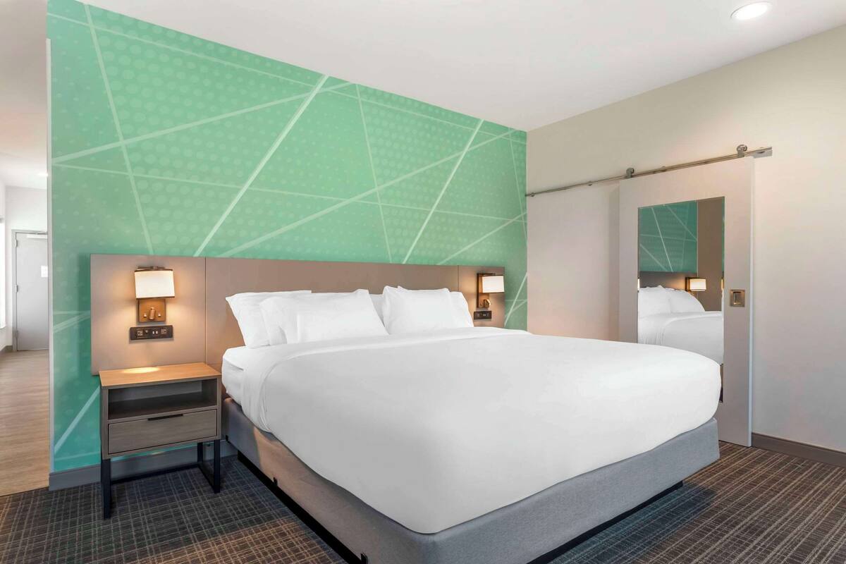 A modern hotel room with a large bed, a nightstand with a lamp, and a mirror on the wall.