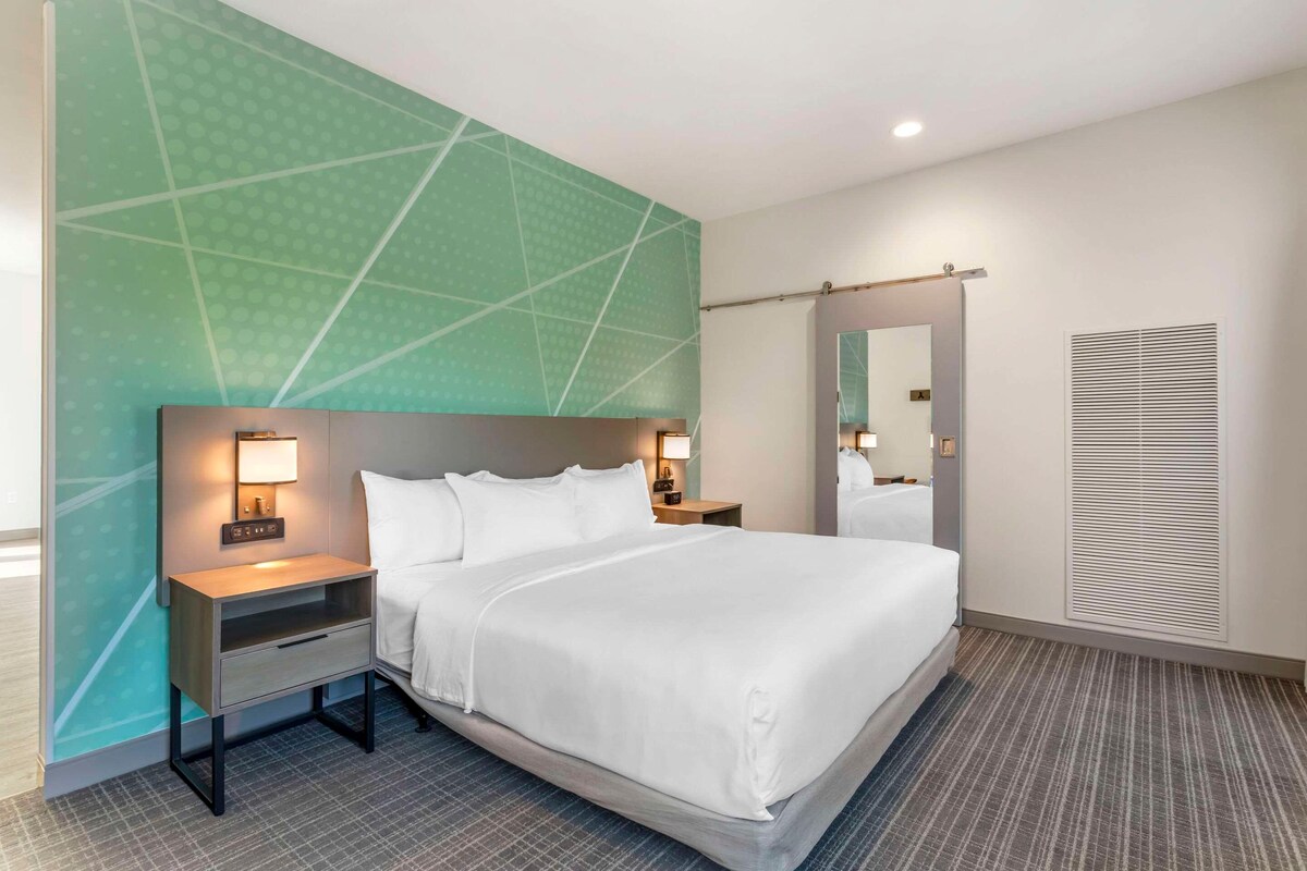 A hotel room with a large bed, two bedside tables with lamps, a mirror, and a window with blinds.