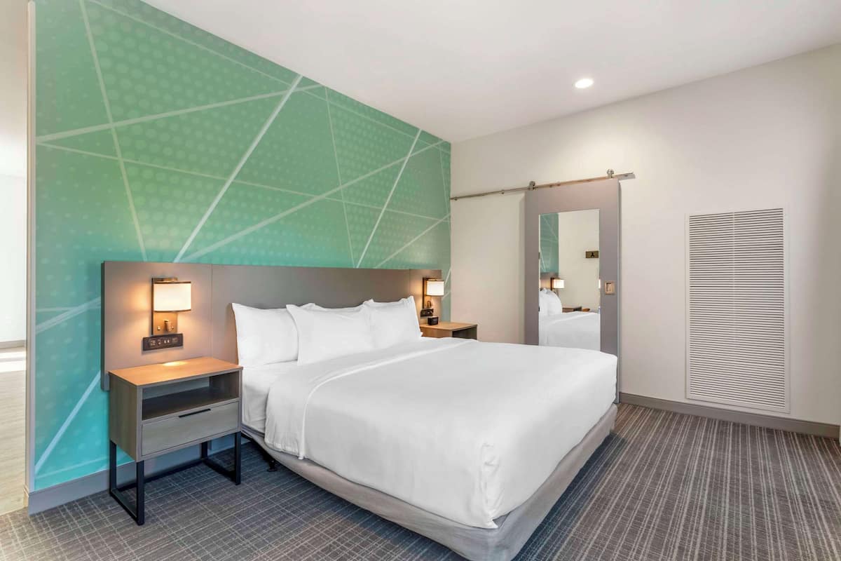A hotel room with a large bed, a nightstand with a lamp, a mirror, and a wall with a geometric pattern.