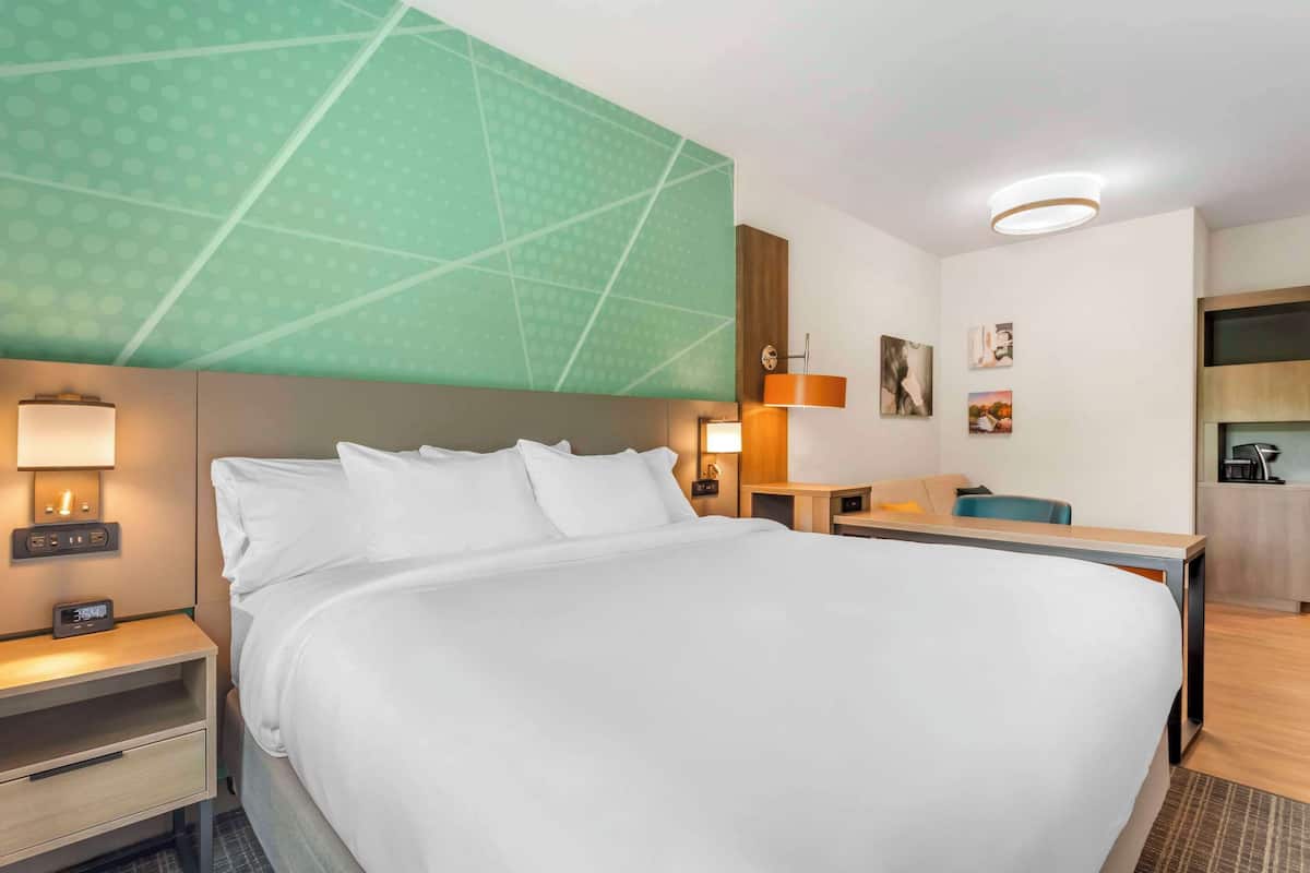 A hotel room with a large bed, a desk, a chair, a nightstand, a wall clock, and a painting on the wall.