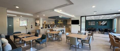 A modern dining area with tables and chairs, a kitchen in the background, and a large window.