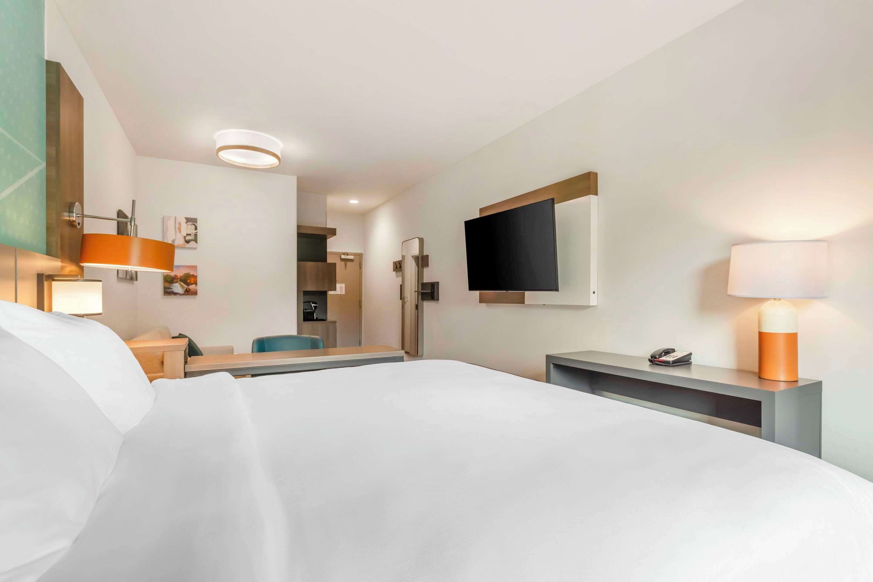 Une chambre d’hôtel avec un grand lit, un bureau avec une lampe, un téléviseur fixé au mur et une salle de bain visible à travers une porte ouverte.