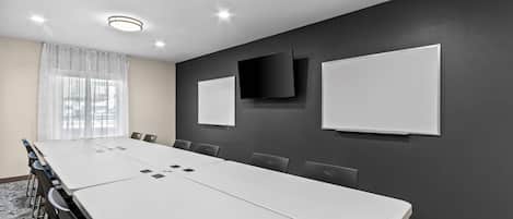 A conference room with a long table, chairs, a whiteboard, a TV, and a window with curtains.