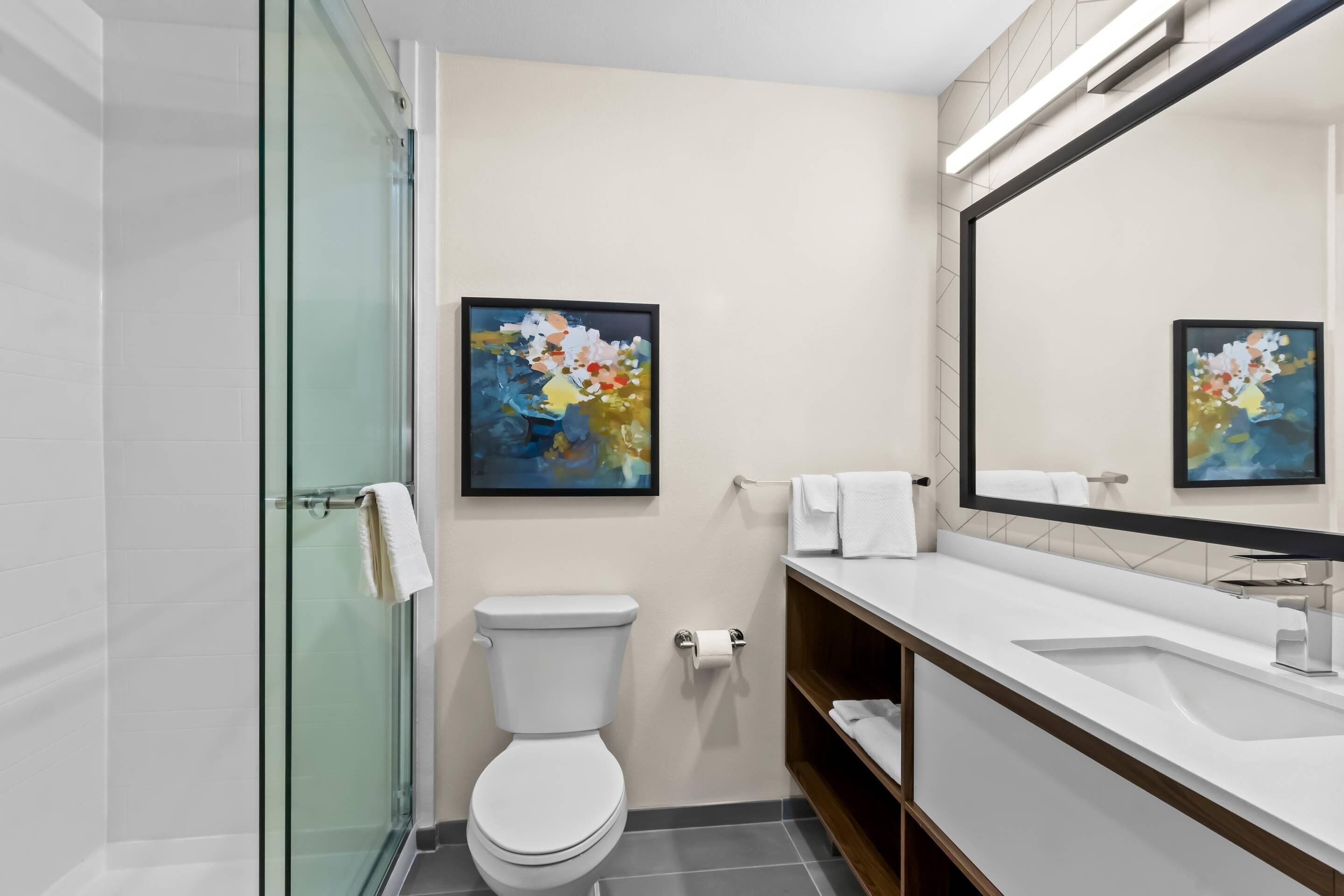 Un baño moderno con cabina de ducha de vidrio, inodoro blanco, lavamanos blanco con un espejo grande y dos cuadros abstractos enmarcados en la pared.