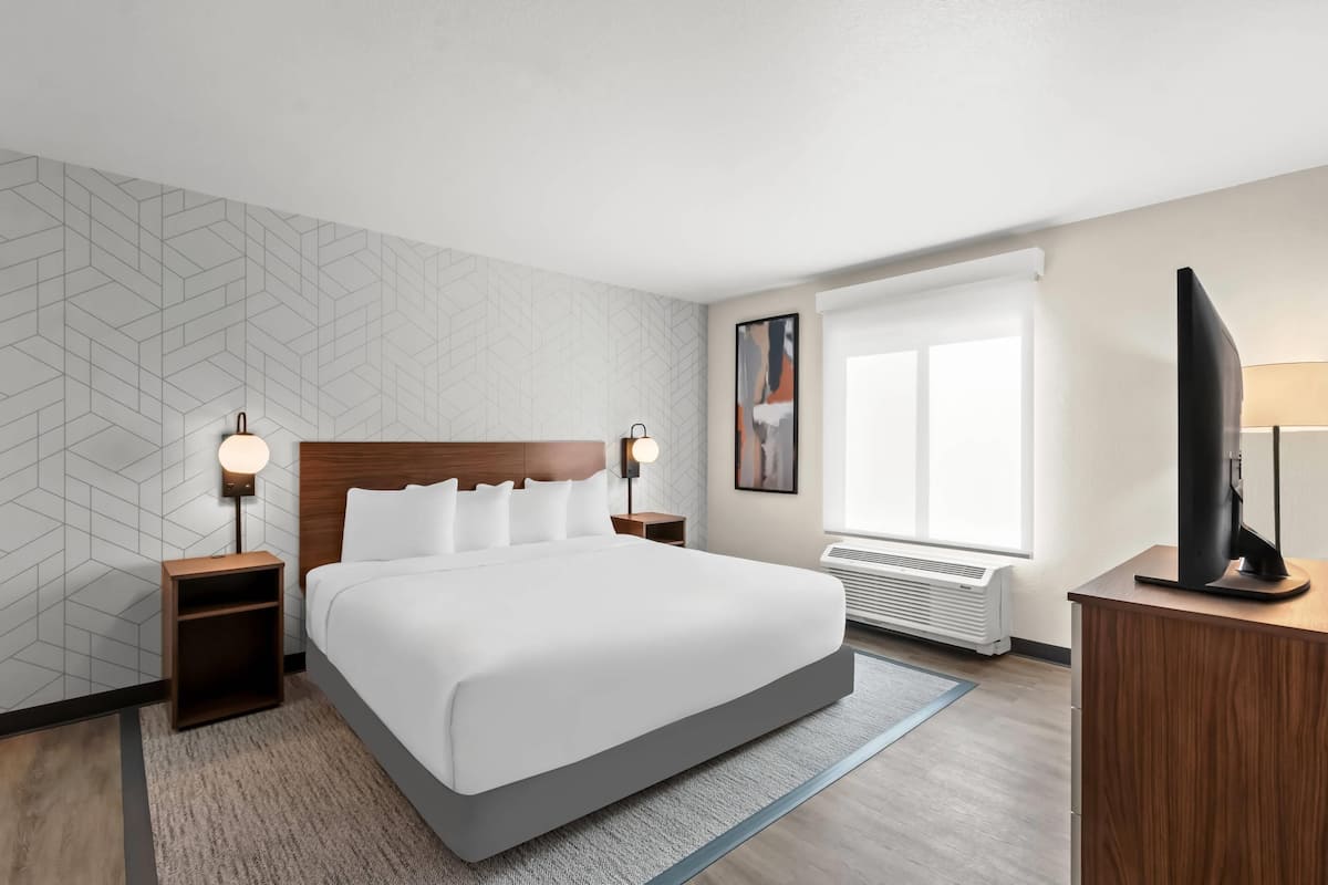 Una habitación de hotel moderna con una cama grande, dos mesitas de noche con lámparas, un televisor de pantalla plana, una ventana con persianas y un aire acondicionado instalado en la pared.