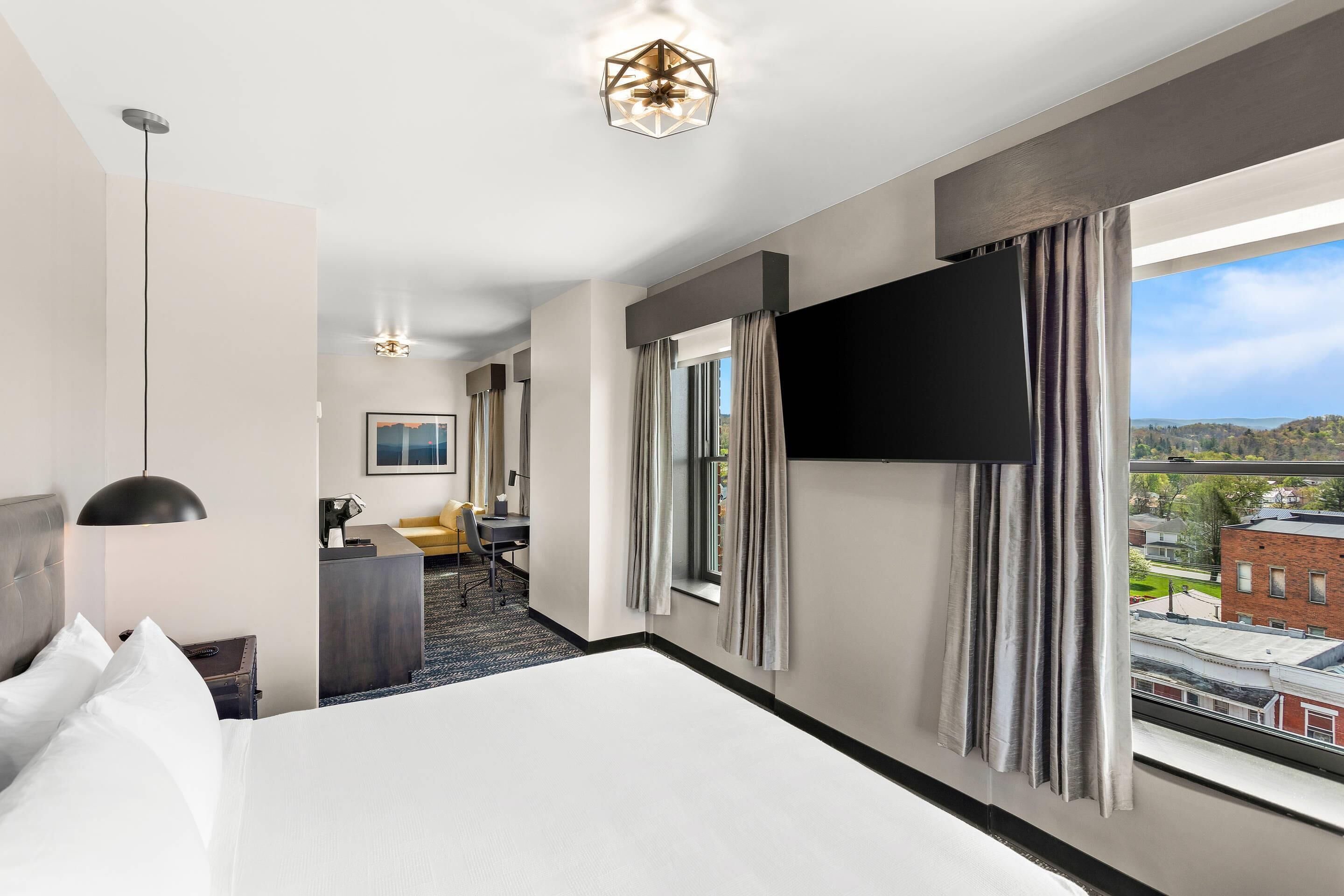 A hotel room with a large bed, a desk with a chair, a flat-screen TV mounted on the wall, and a view of the city through the window.