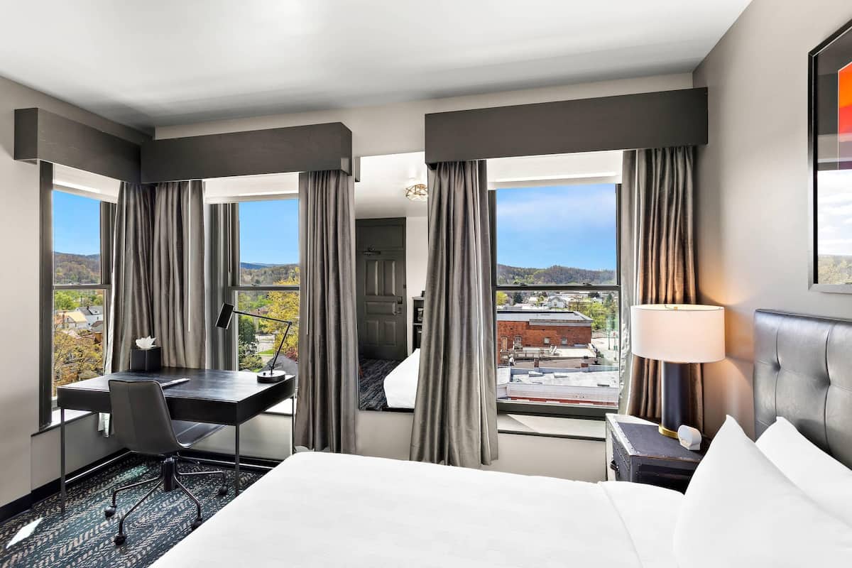 A hotel room with a bed, desk, and chair, large windows with curtains, a nightstand with a lamp, and a view of the city.