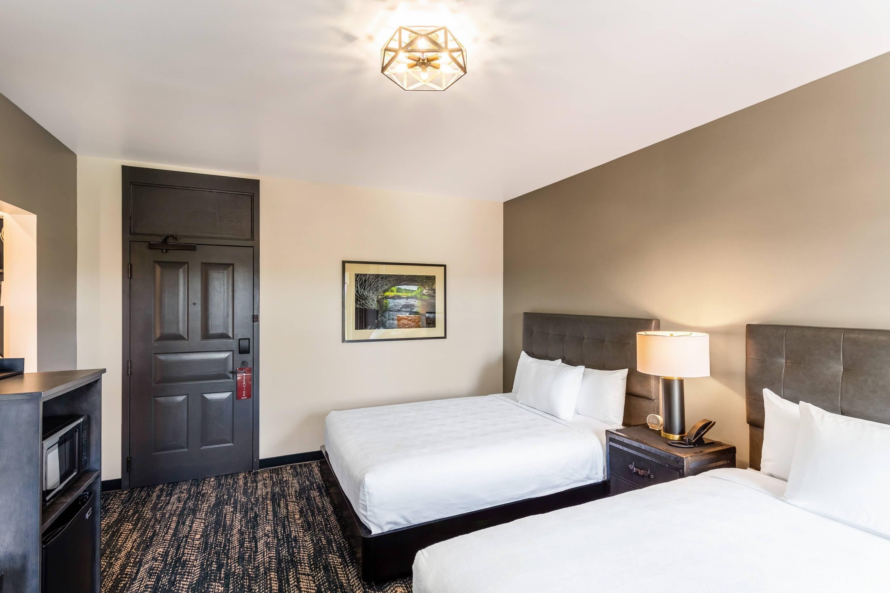 A hotel room with two beds, a desk, a chair, a door, and a ceiling light fixture.