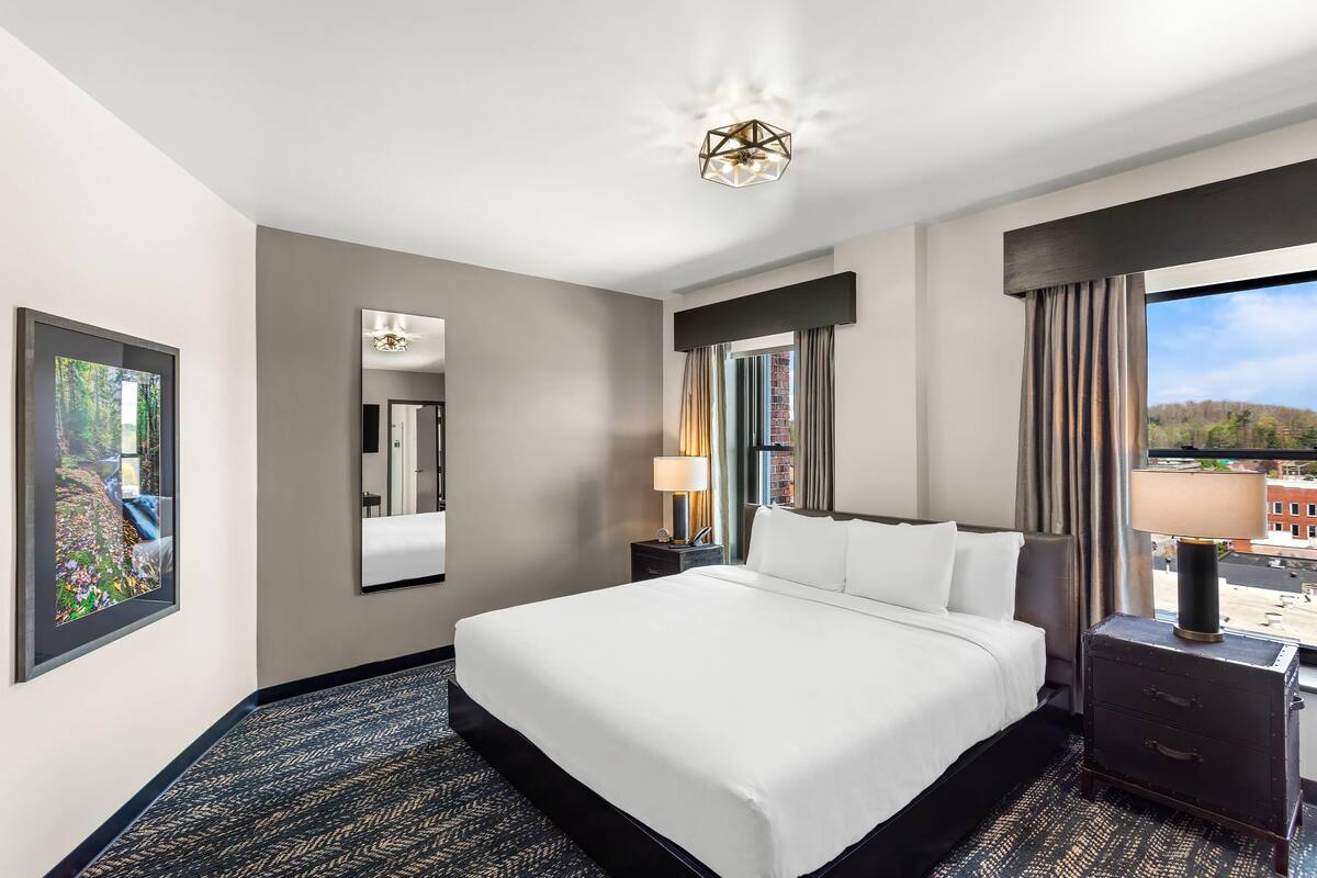 A hotel room with a large bed, a nightstand with a lamp, a dresser, a large mirror, and a window with curtains.