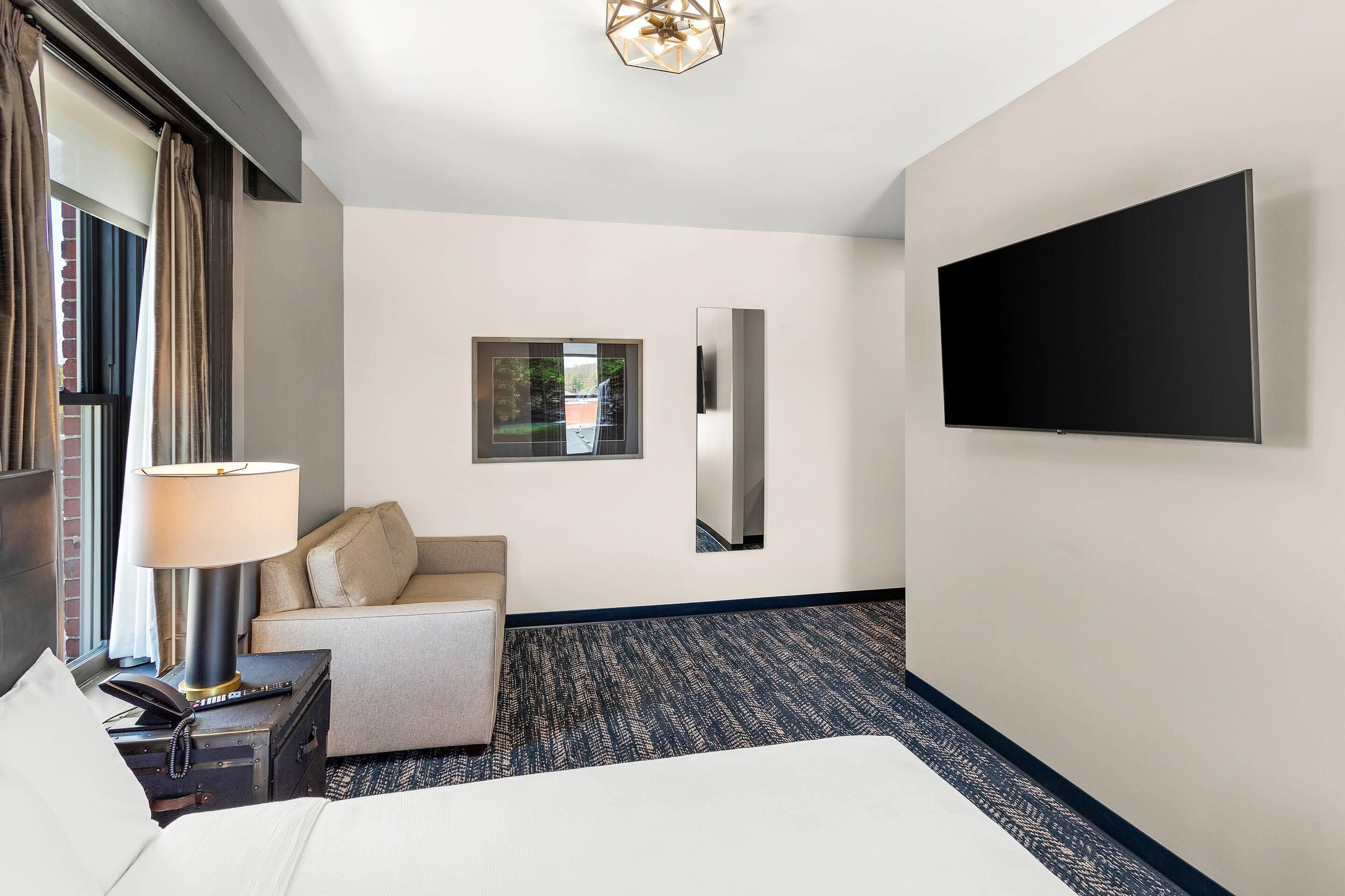Habitación de hotel con cama, escritorio con lámpara, televisor montado en la pared y vistas al exterior a través de una ventana.