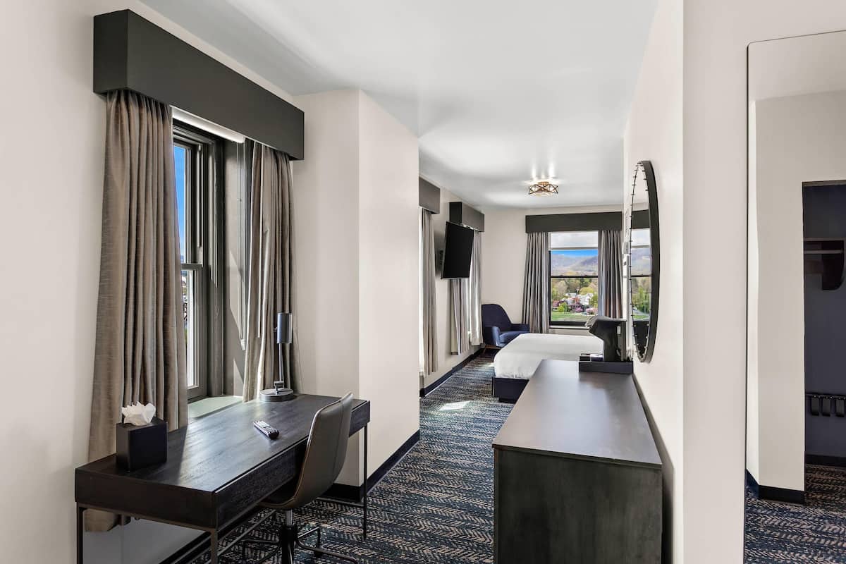 A hotel room with a bed, a desk, a chair, a TV, and a window with curtains.