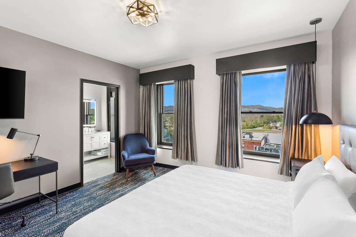 A hotel room with a large bed, a desk with a lamp, a chair, and a view of the outside through large windows.