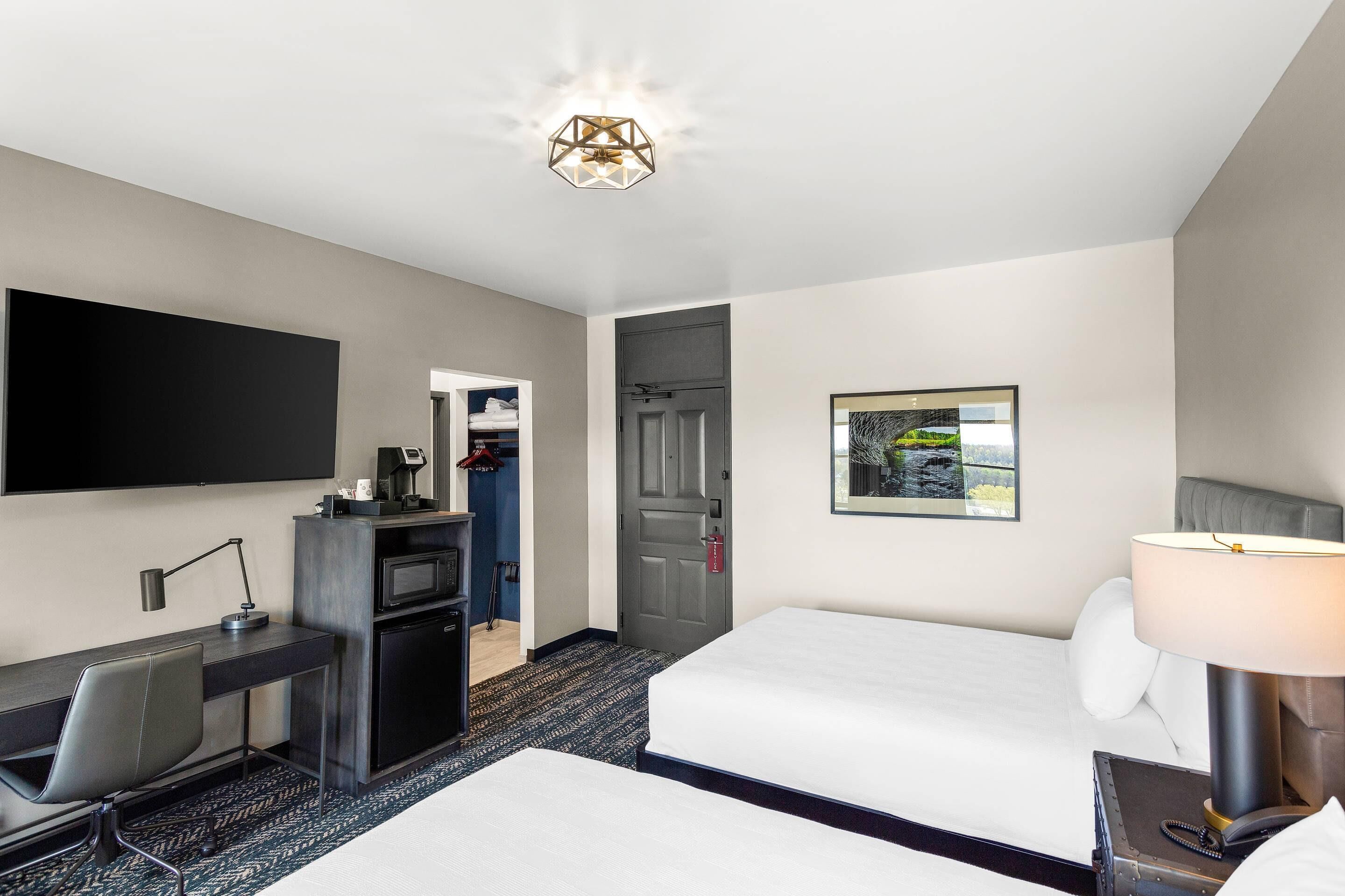 Een hotelkamer met een bureau, twee bedden, een televisie, een bureaulamp, een nachtkastje met lamp, een lijstbeeld aan de muur en een deur naar een kast.
