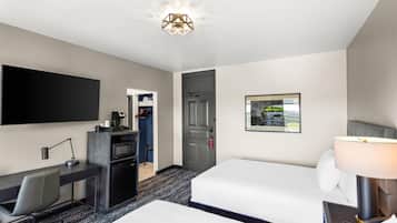 Een hotelkamer met een bureau, twee bedden, een televisie, een bureaulamp, een nachtkastje met lamp, een lijstbeeld aan de muur en een deur naar een kast.