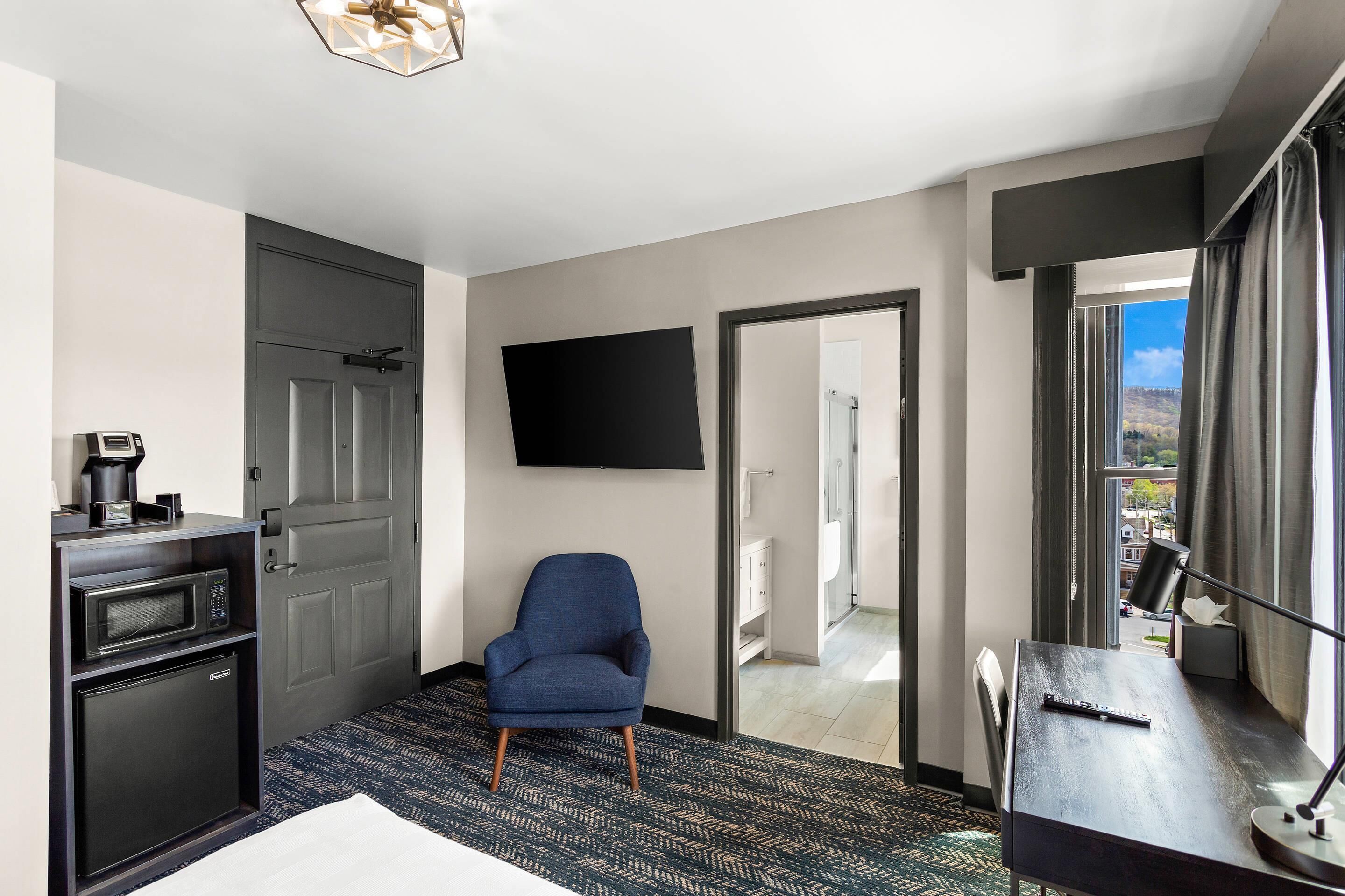 Habitación de hotel con un sillón azul oscuro, un televisor de pantalla plana, una kitchenette pequeña con microondas y refrigerador, y vista de la ciudad a través de un ventanal grande.
